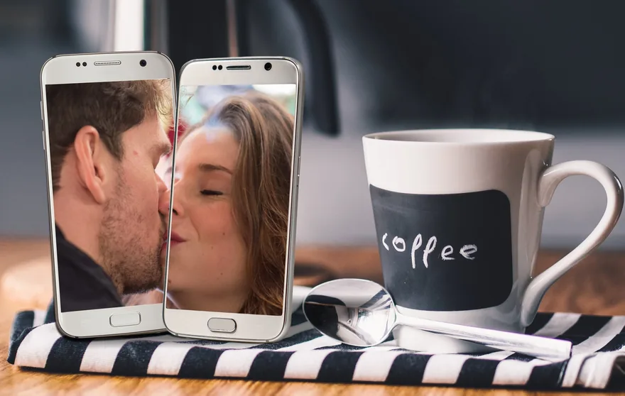 Two phones showing a couple kissing on a video call, beside a coffee mug